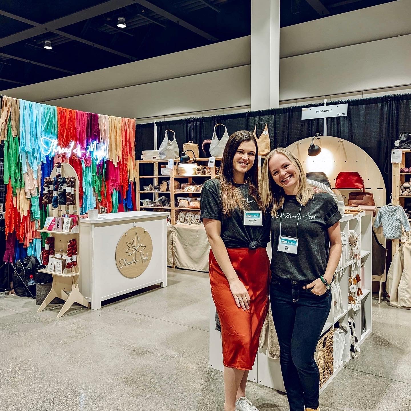 Two women smiling at a trade show booth featuring colorful fabrics and handmade bags.