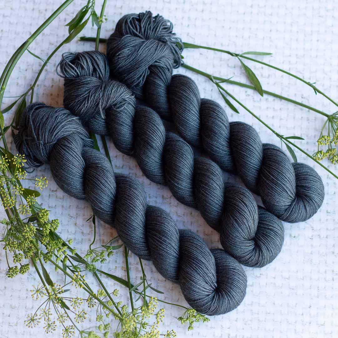 Three skeins of dark gray yarn with greenery on a textured white background.