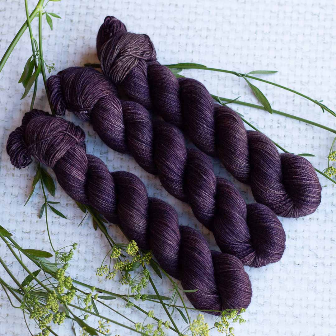 Three skeins of dark purple yarn with green plant accents on a textured white background.