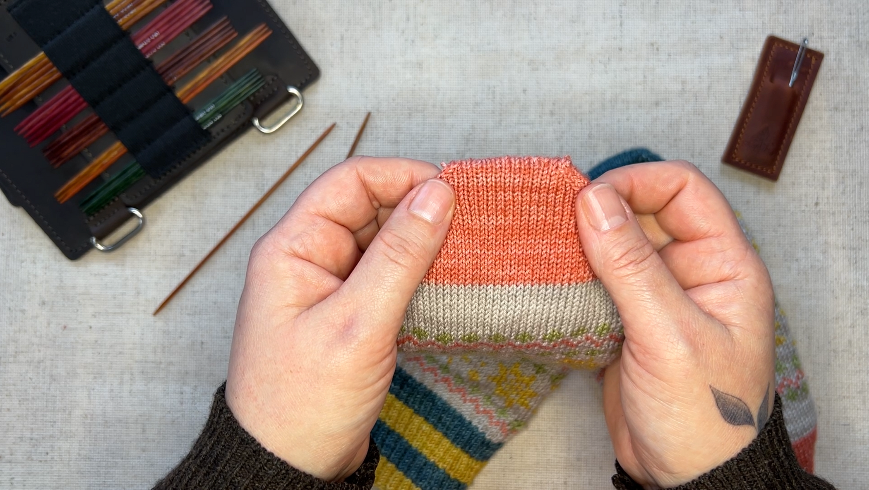 A pair of hands holding the toe of a pink, knitted sock. There are double pointed needles, a tapestry needle in it's sheath, and a Thread & Maple double pointed needle page on the table.