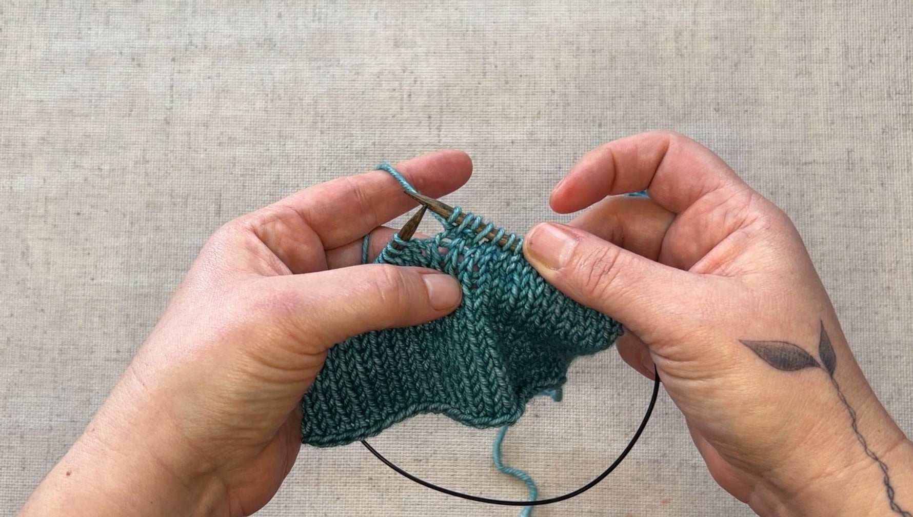 A pair of hands holding circular knitting needles with teal yarn.