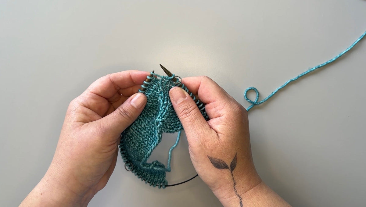Hands are holding circular knitting needles with a piece of turquoise knitted fabric on the needles.