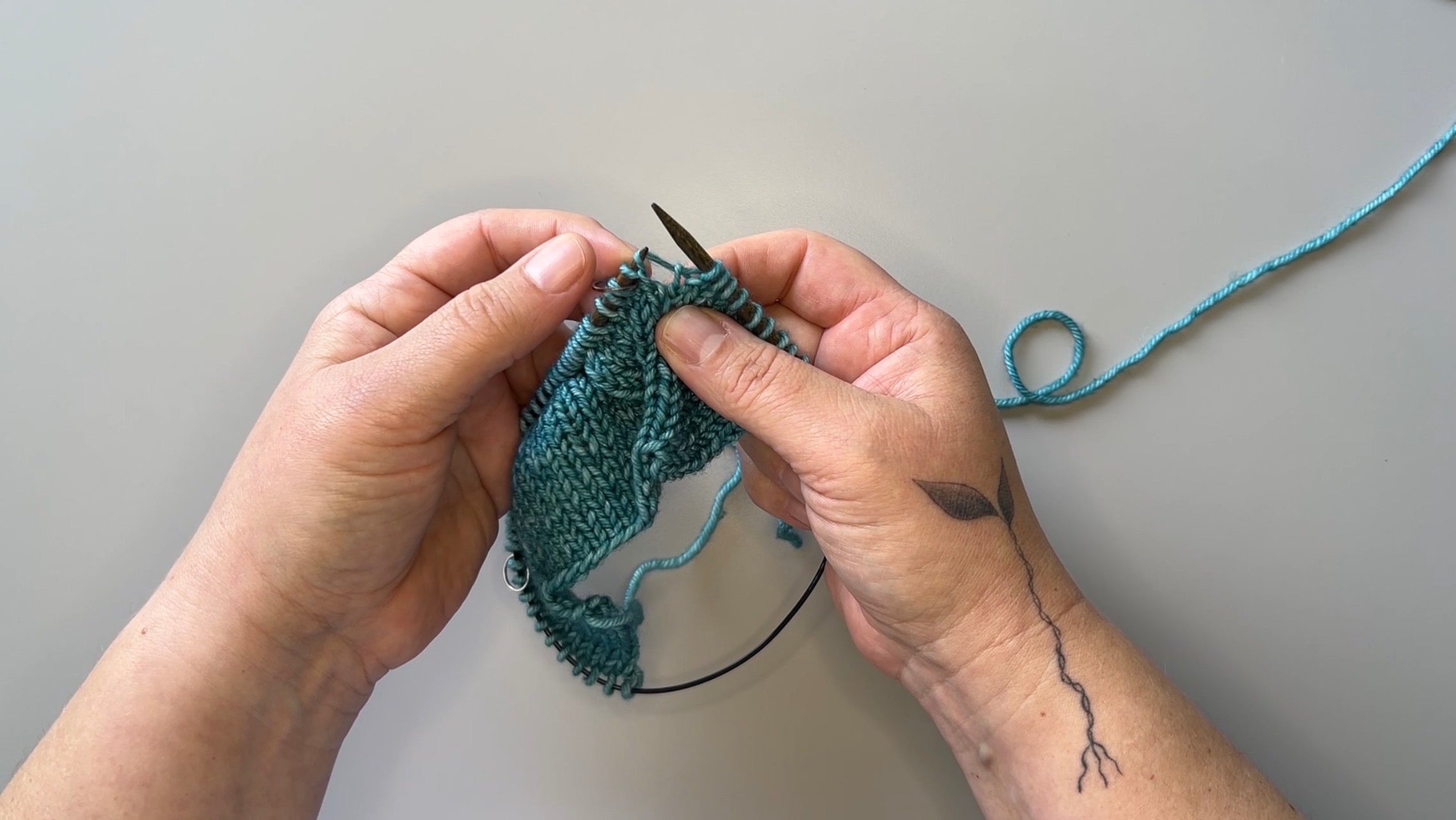 Hands holding circular knitting needles with turquoise knitted fabric on the needles.