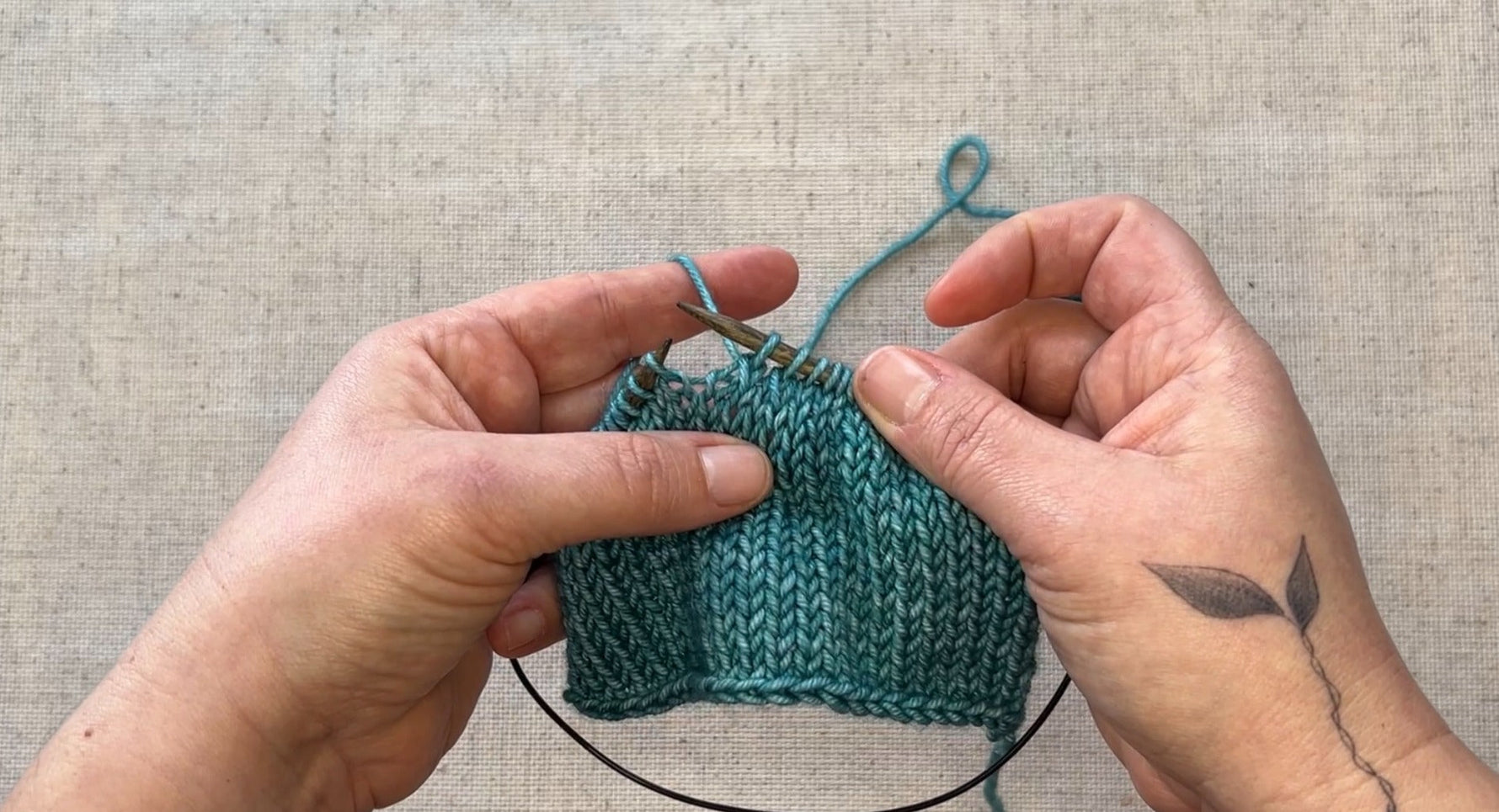A pair of hands is holding circular knitting needles and a swatch in teal yarn. 
