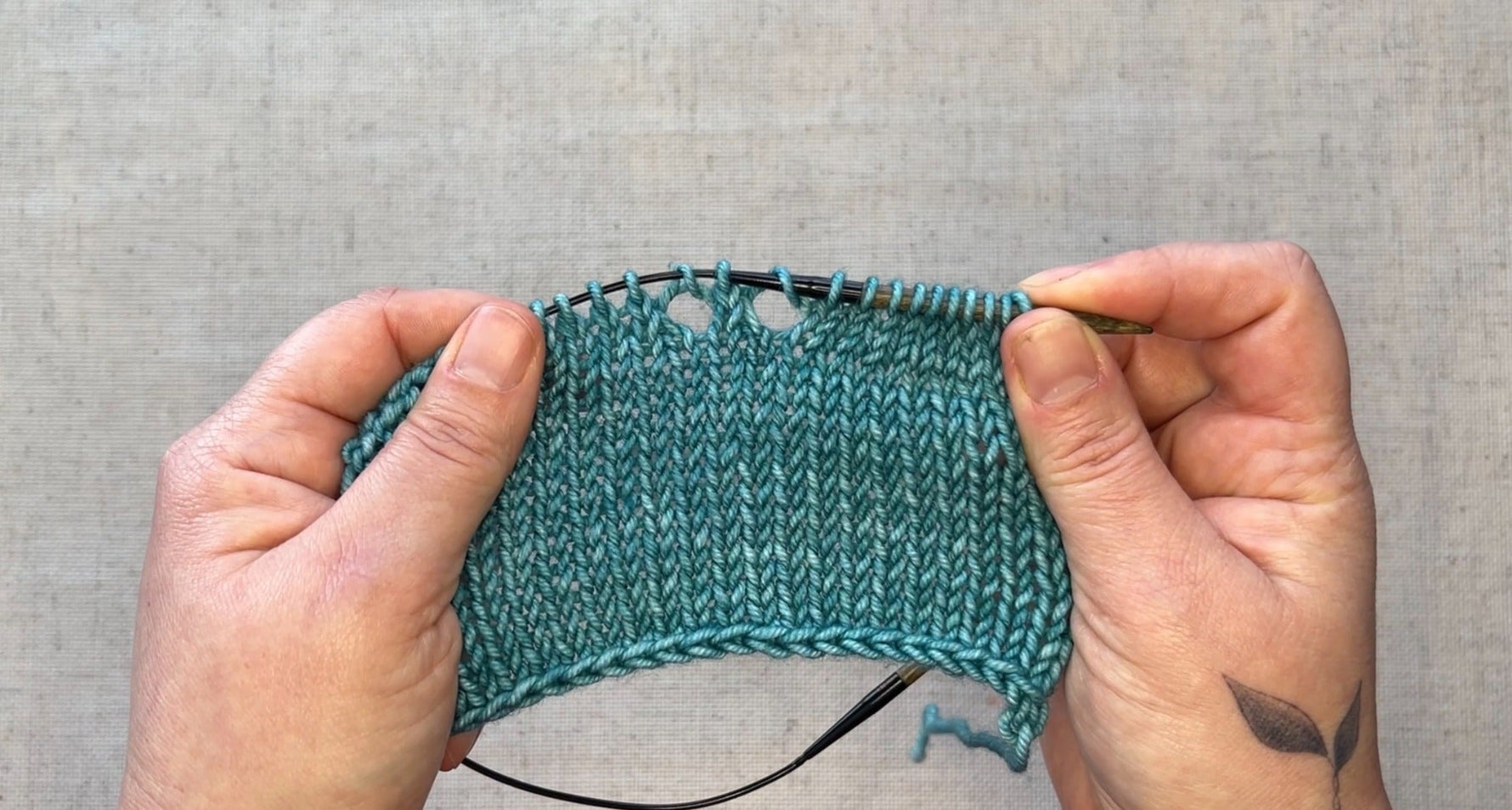 Two hands hold a piece of teal knitted fabric with two holes at the top, demonstrating what a yarn over looks like. 