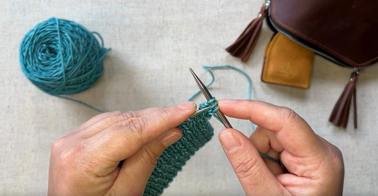 Hands knitting with turquoise yarn and bag.