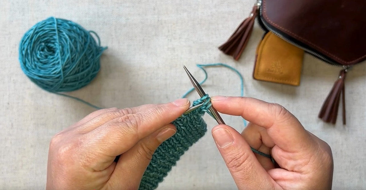 Hands knitting with turquoise yarn and bag.