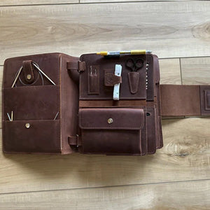 Open brown leather portfolio organizer on wooden floor with pockets, pens, sticky notes, and scissors.