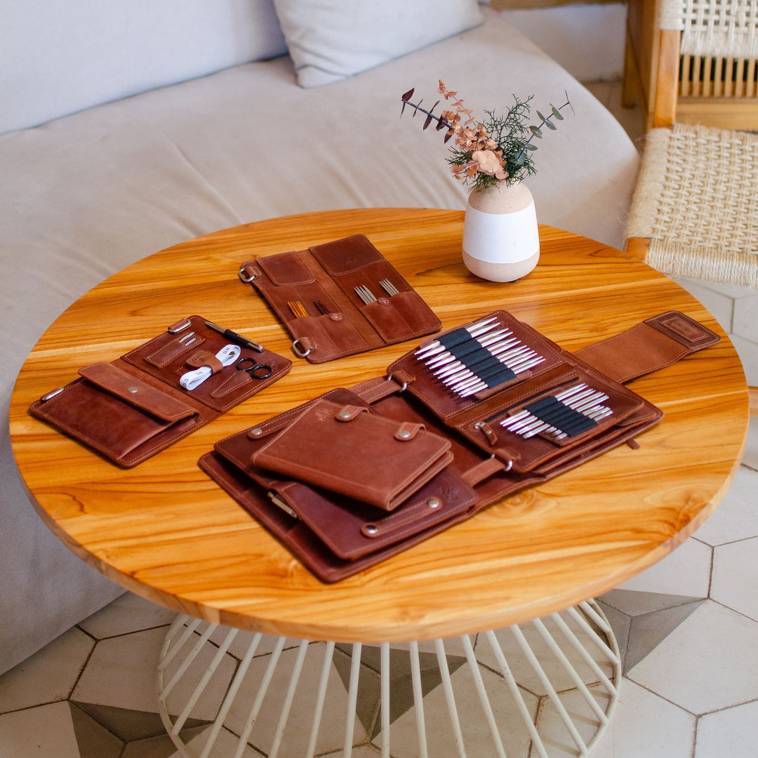 Brown leather travel accessories neatly arranged on a wooden round table with a white vase and plant.